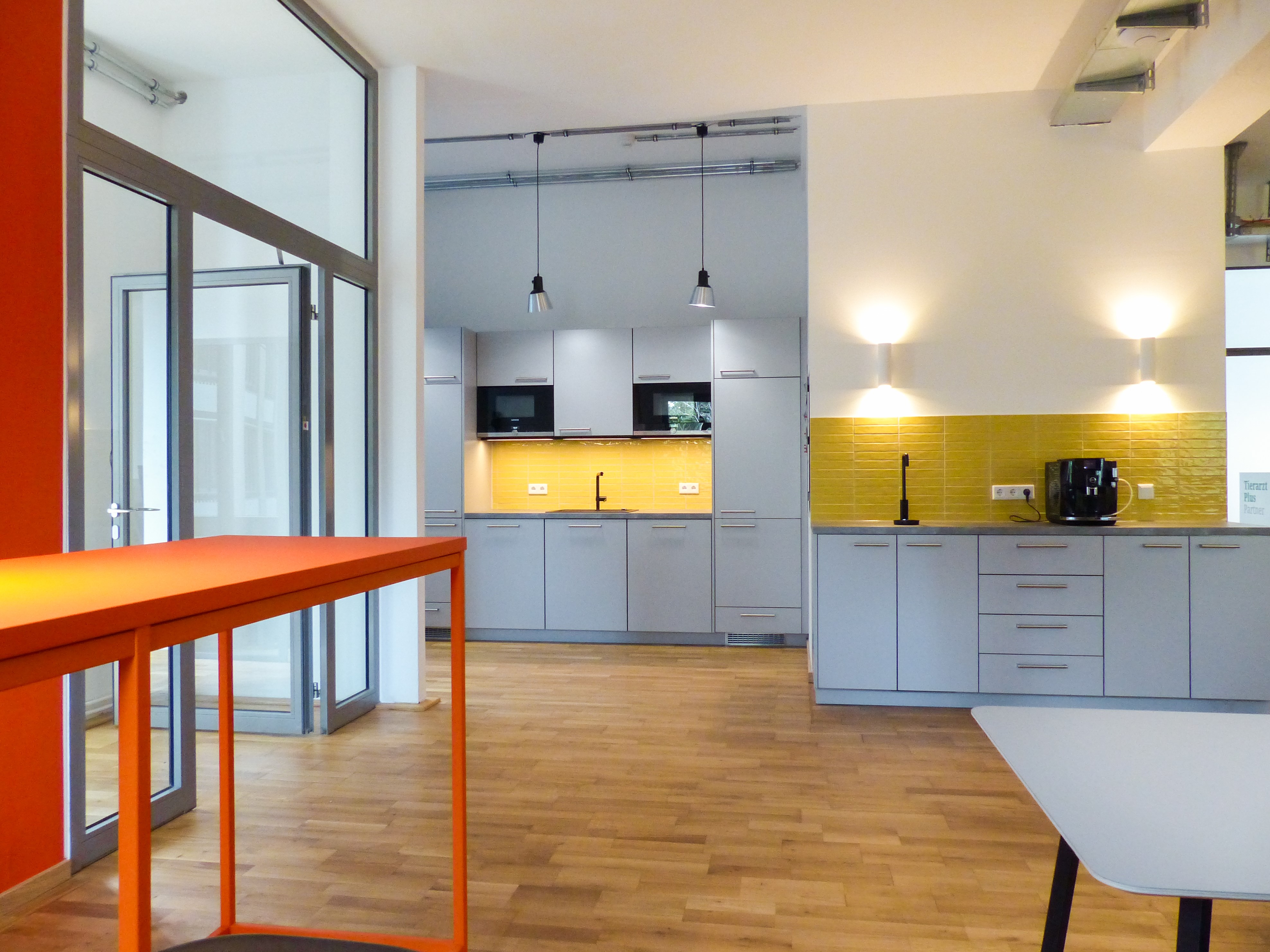 Moderne graue Küchenzeile mit gelber Fliesenrückwand und Holzfußboden, Teil einer Tierarztpraxis mit minimalistischen Pendelleuchten und oranger Akzentwand.